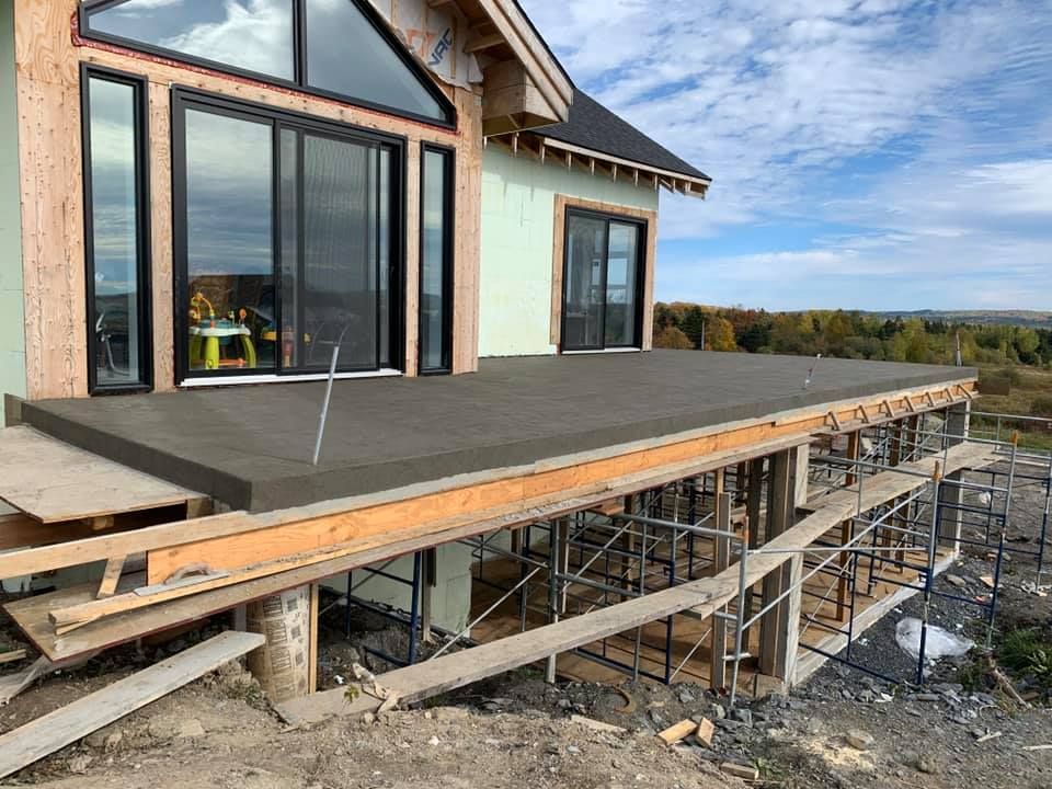 Terrasse en béton en construction, attachée à une maison avec de grandes fenêtres, échafaudage en dessous.
