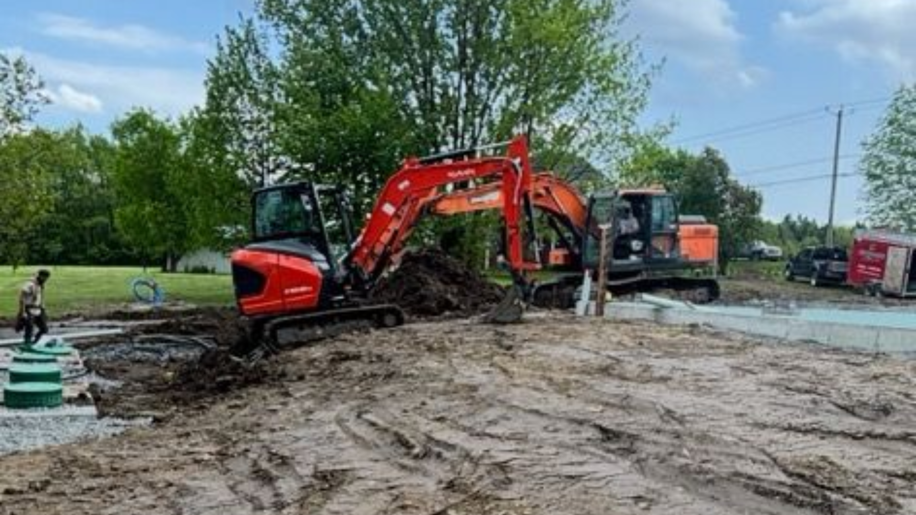 Deux excavatrices creusent sur un chantier. L'une est orange, l'autre noire. Sol boueux et arbres verts.