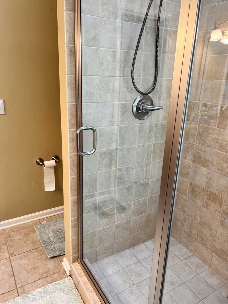 Shower stall with glass doors, tiled walls, and showerhead. Beside it is a wall with a toilet paper holder.