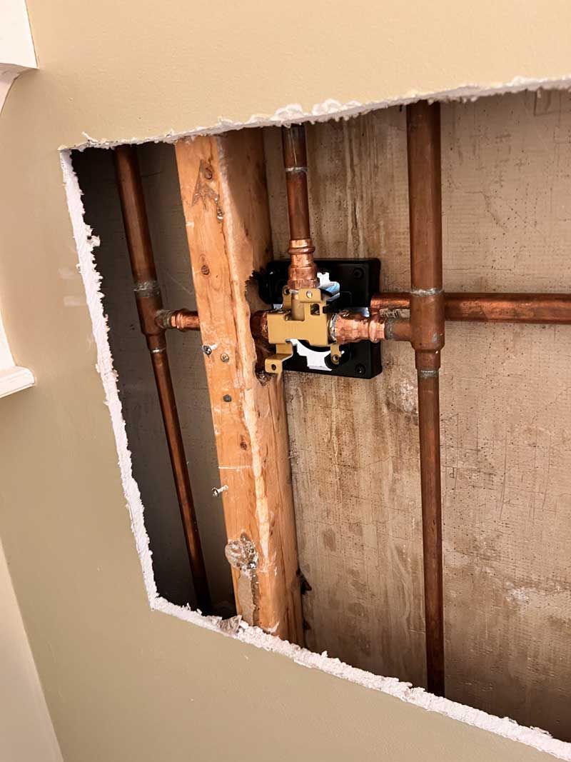 Copper plumbing inside a wall, including a shower valve, exposed by a hole cut into the drywall.