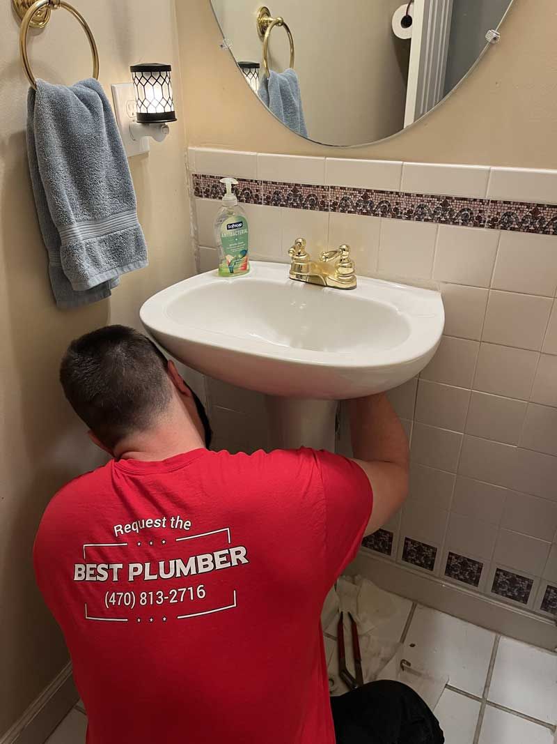 Plumber in red shirt working on a pedestal sink in a bathroom with a mirror.
