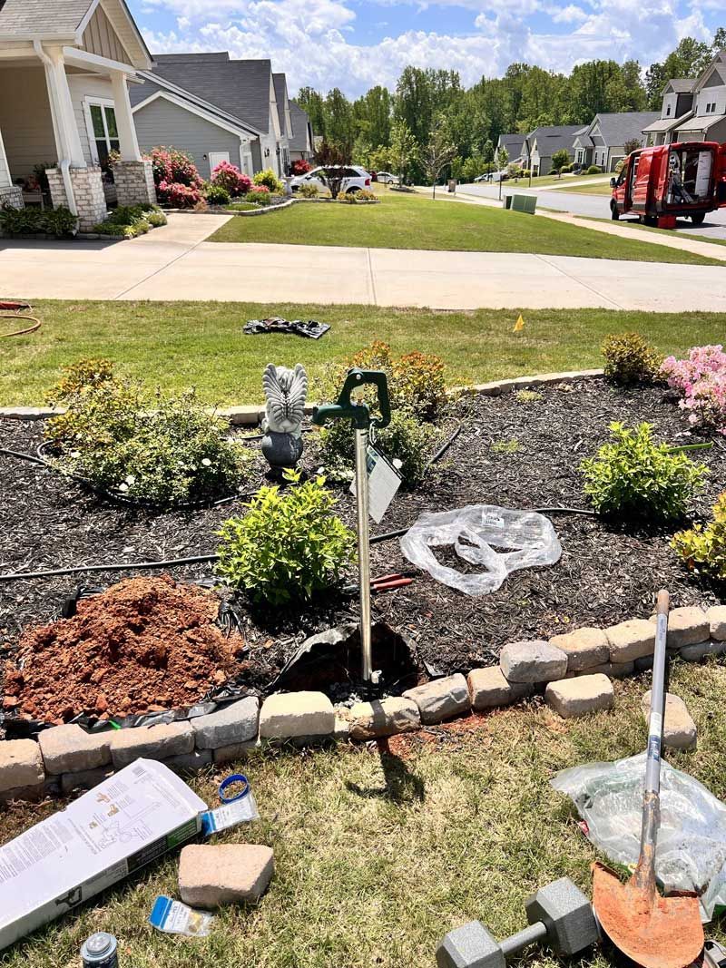Flower bed with sprinkler, mulch, rocks, and tools in front of a house on a sunny day.