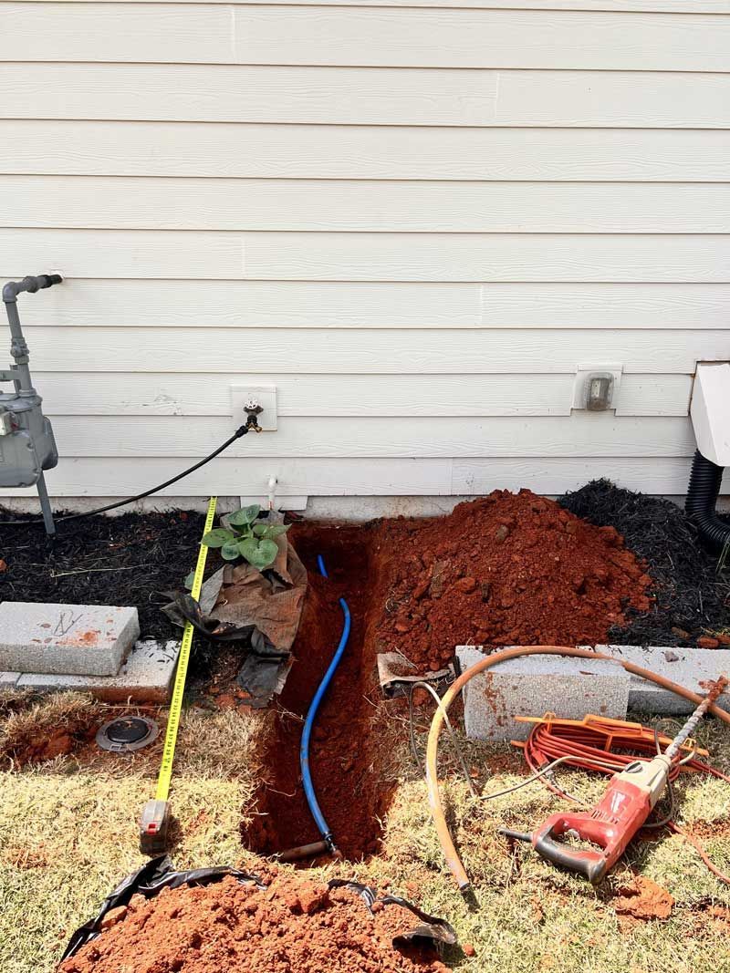Trench dug along house wall with blue pipe visible. Tools and red dirt pile.