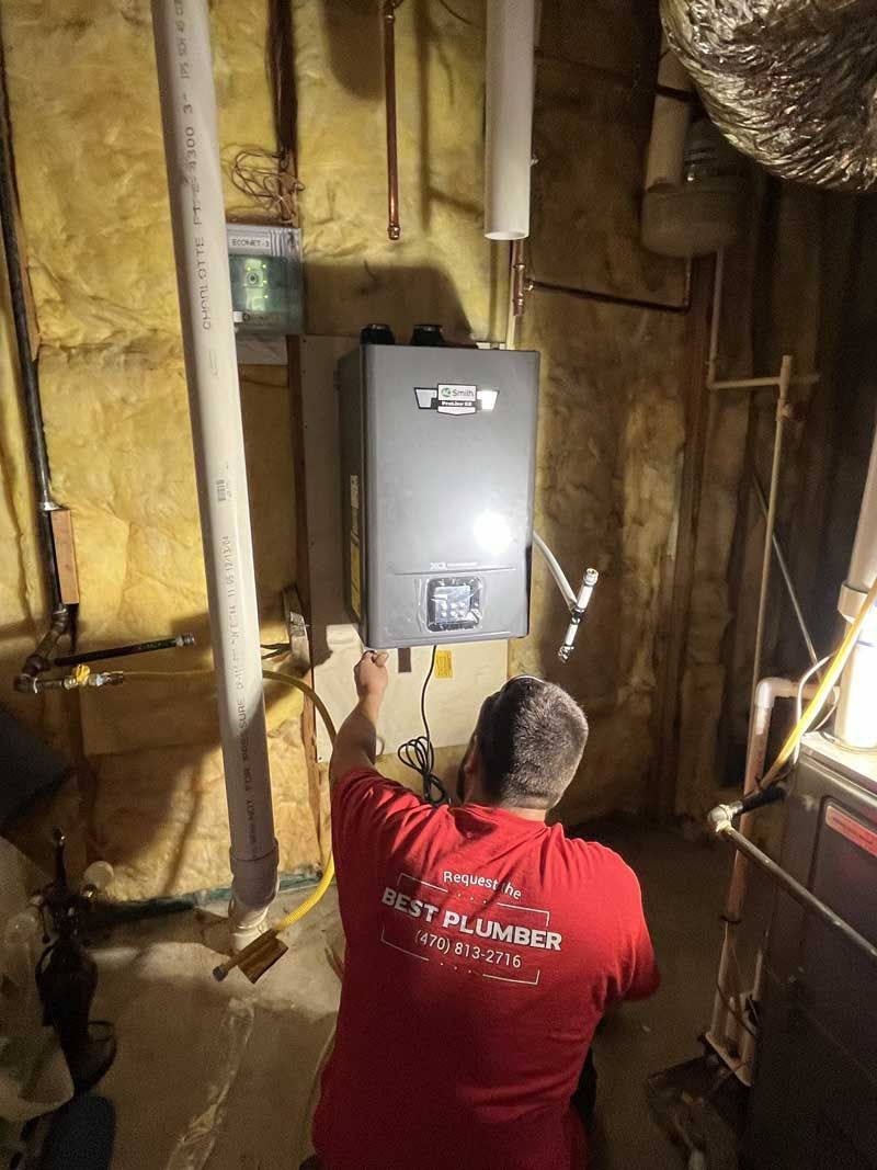 Plumber in red shirt working on a gray tankless water heater in a basement.
