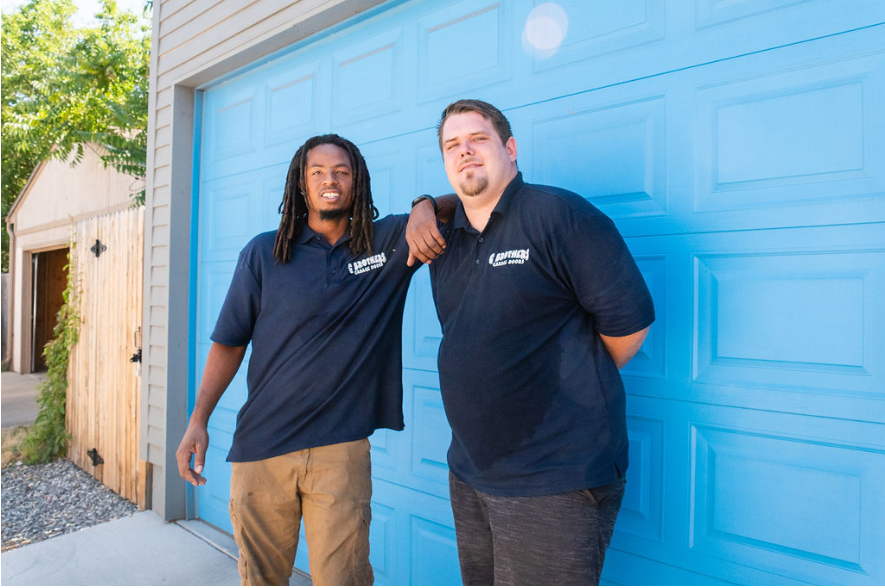 Two men are standing in front of a blue garage door.