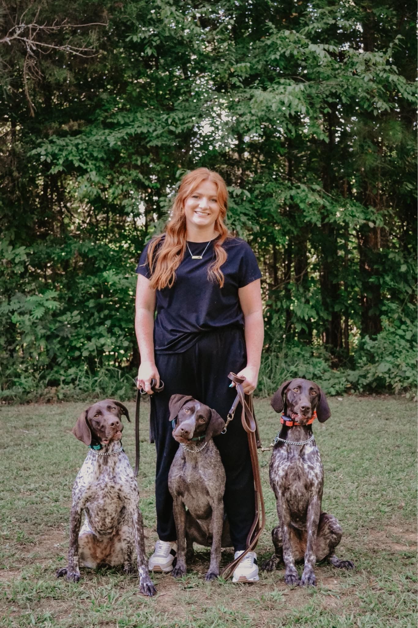 A woman is standing next to three dogs on leashes in a field.