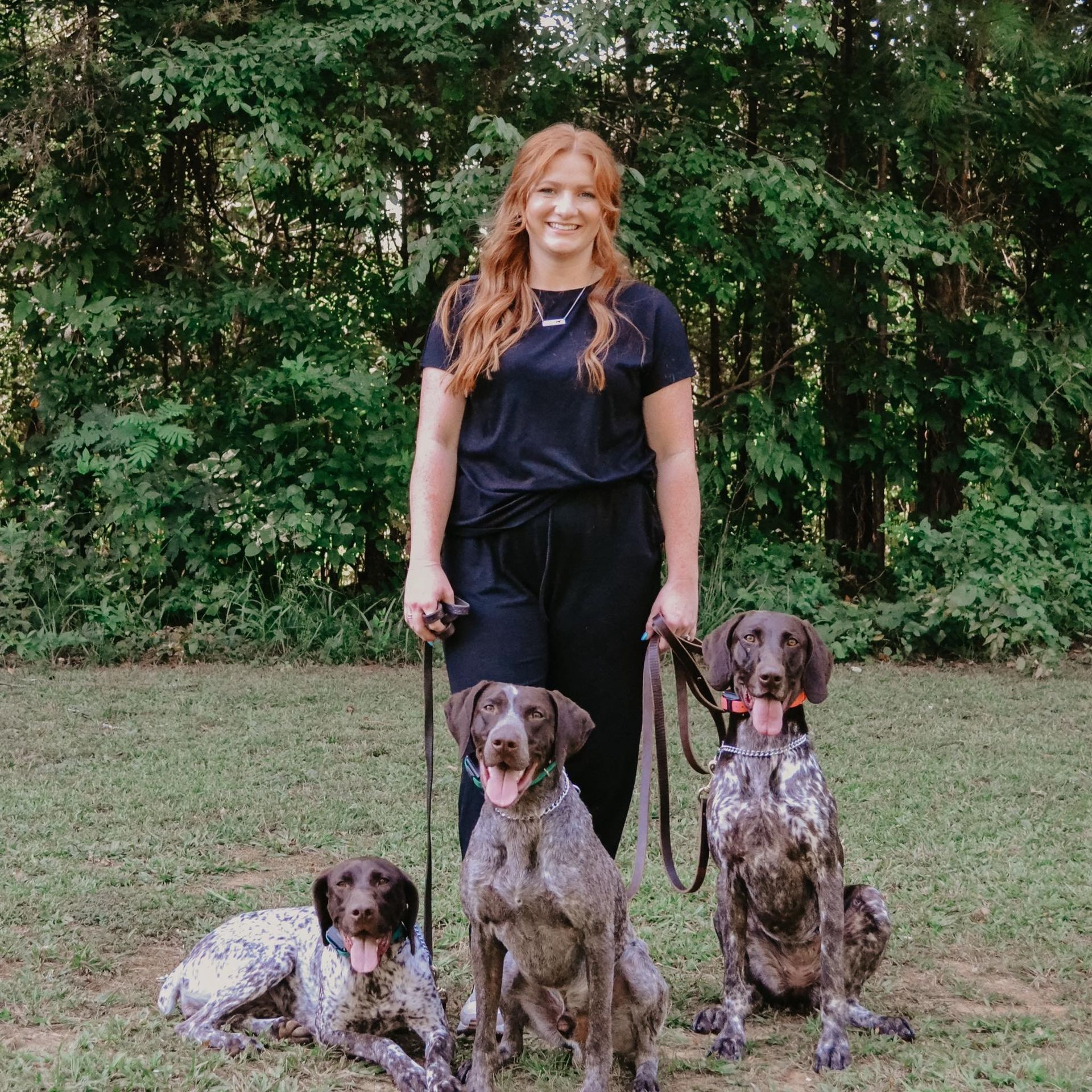 A woman is standing next to three dogs on leashes in a field.