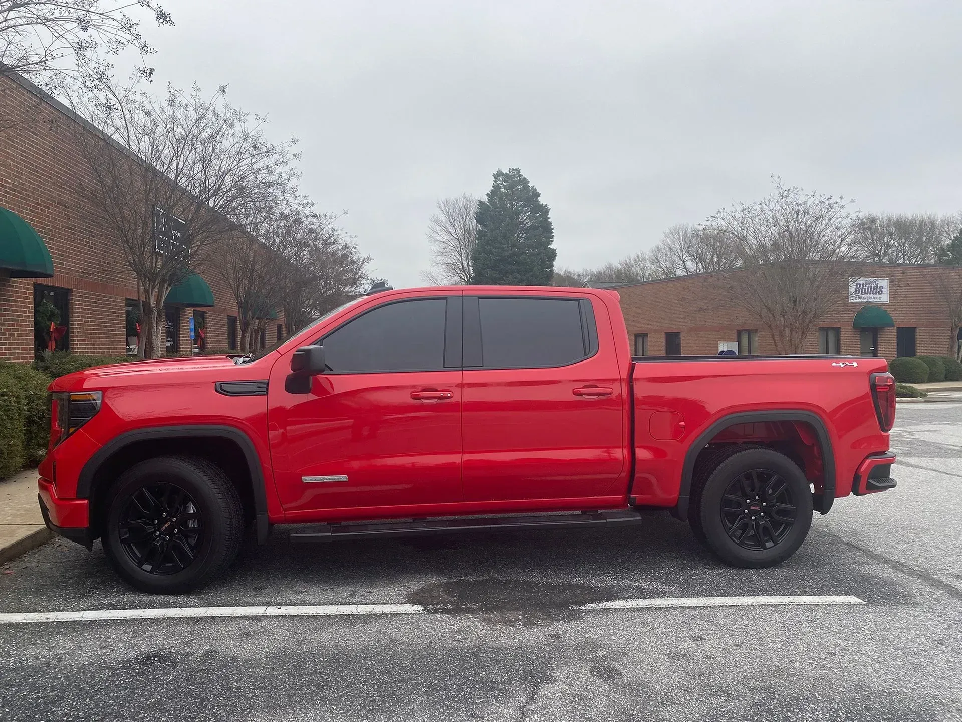 Red pickup truck with black wheels parked in a lot in front of a building on an overcast day.