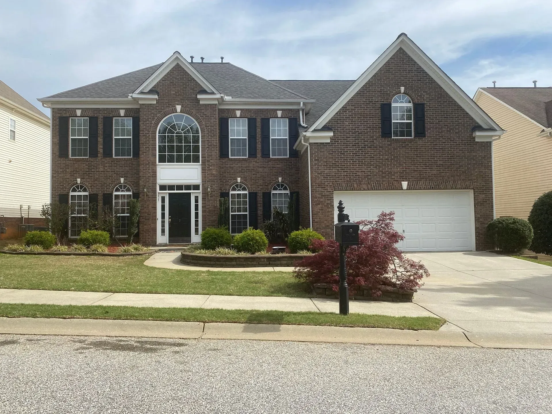 Two-story brick house with black shutters, white garage door, and landscaped front yard.