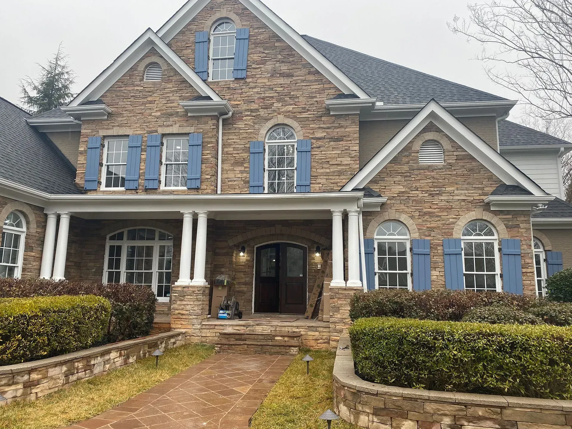 Stone house with blue shutters, front porch, and manicured bushes. Cloudy day.