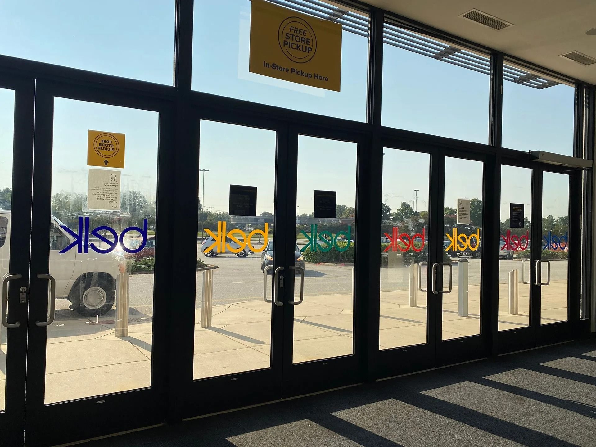 Entrance to Belk store with large glass doors, colorful text, and signs, reflecting the outdoors.