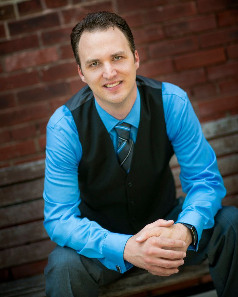 A man in a blue shirt and black vest is sitting on a bench with his hands folded