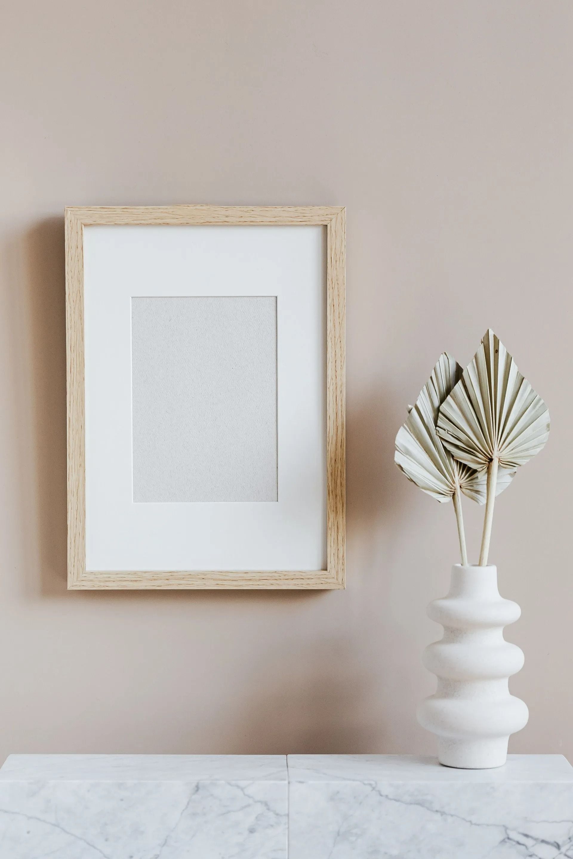 A light wood frame with a white mat hangs on a beige wall next to a white textured vase holding dried palm spears.