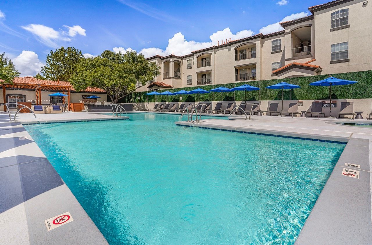 Outdoor pool with lounge chairs and blue umbrellas in a courtyard of an apartment community.