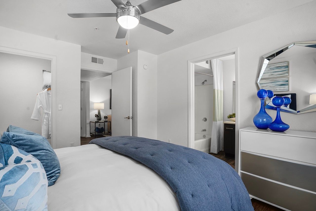 Bedroom in apartment with blue bedspread, dresser, mirror, and door to the bathroom.