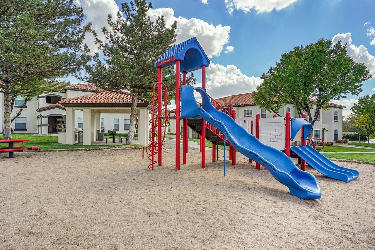 Playground with blue slides and red climbing structures in a sandy area at a residential complex.