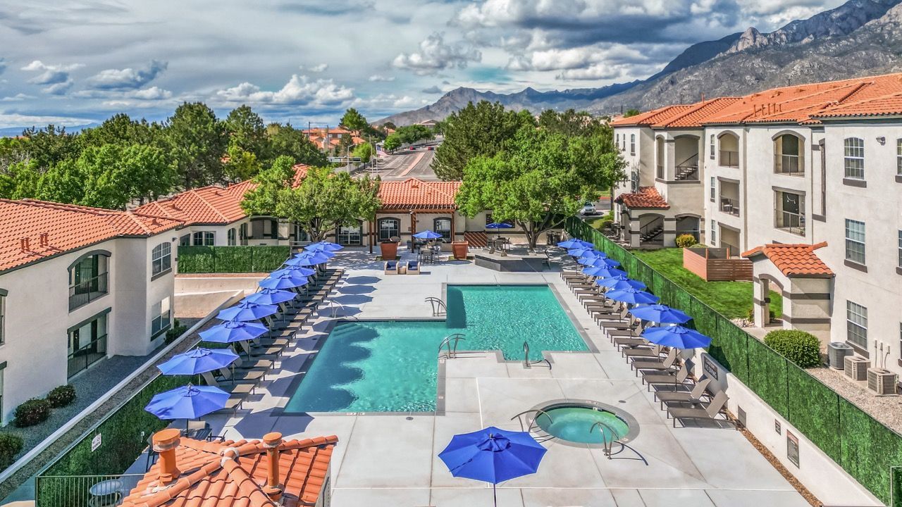 Aerial view of apartment pool deck with blue umbrellas, lounge chairs, and a circular hot tub.