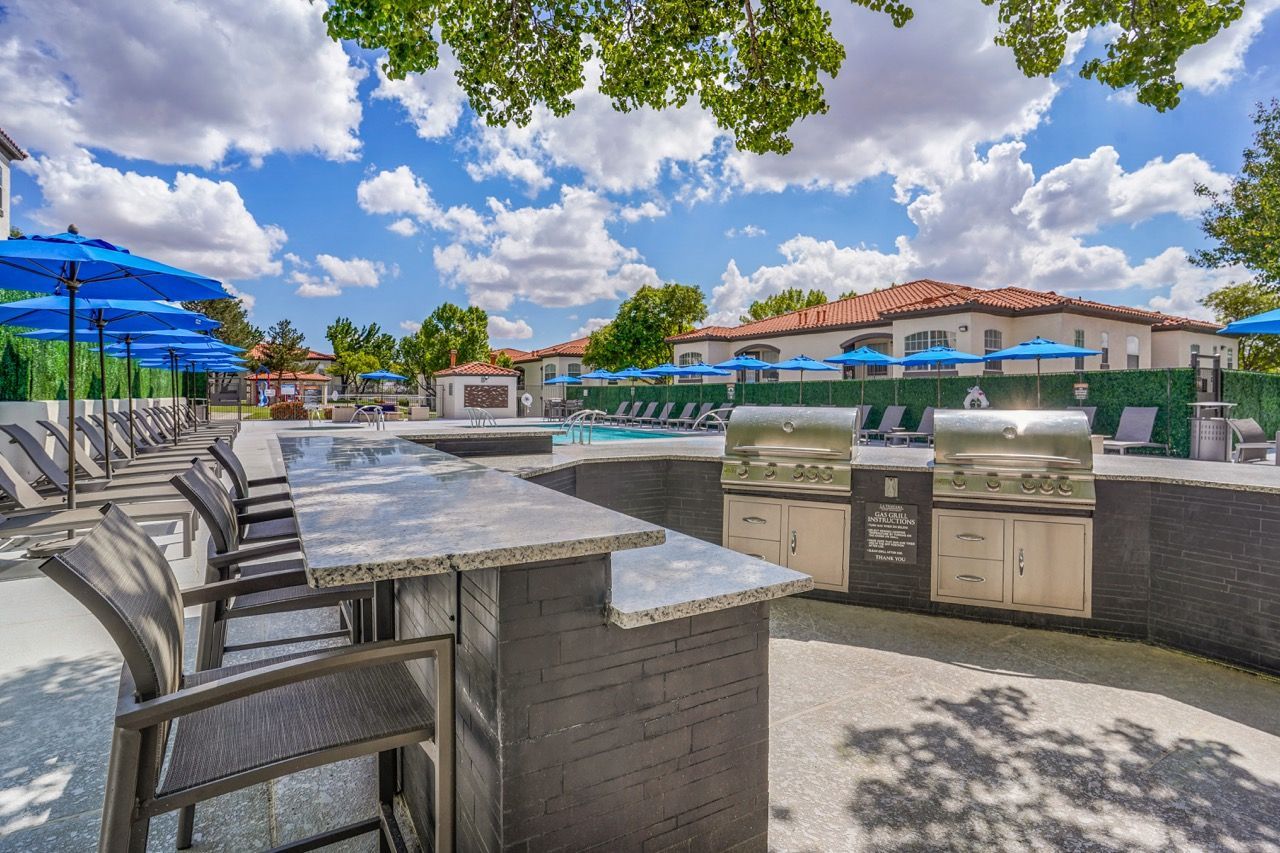 Outdoor apartment community pool area with grilling stations, lounge chairs, and blue umbrellas.
