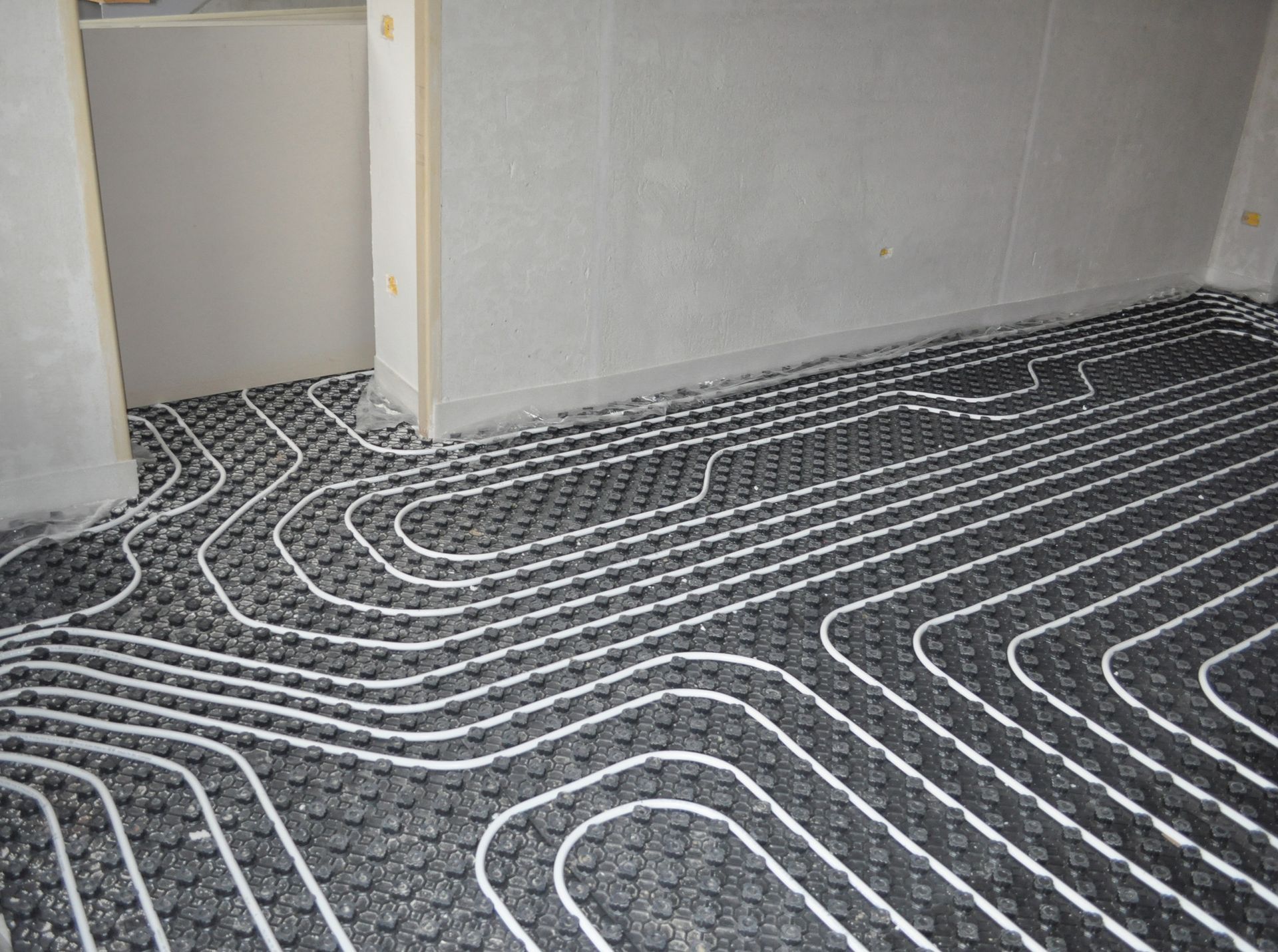 Impianto di riscaldamento a pavimento con tubi bianchi posati su una base nera in una stanza non rifinita.