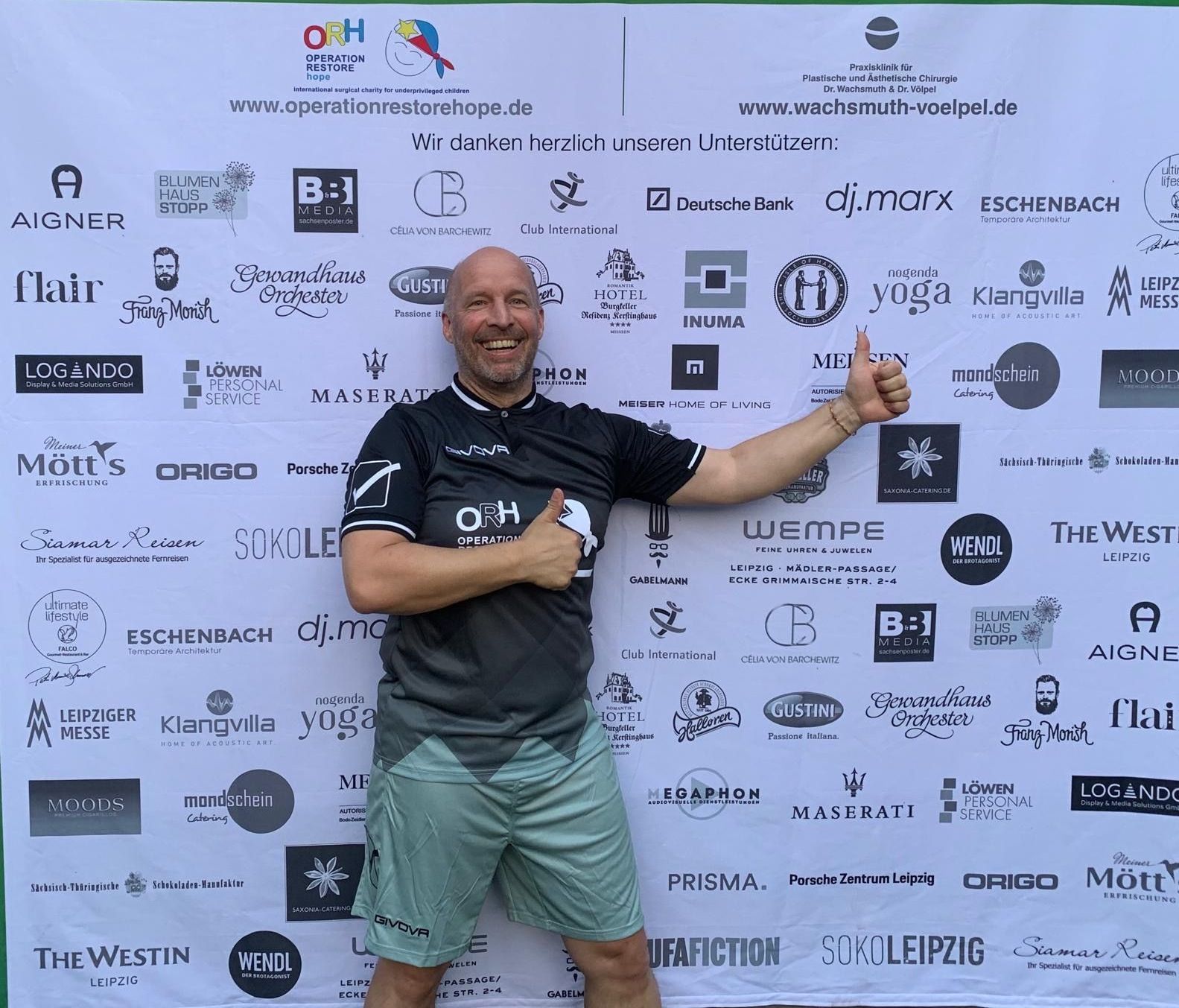 Ein Mann, der vor einer Wand mit Logos einen Daumen hoch zeigt