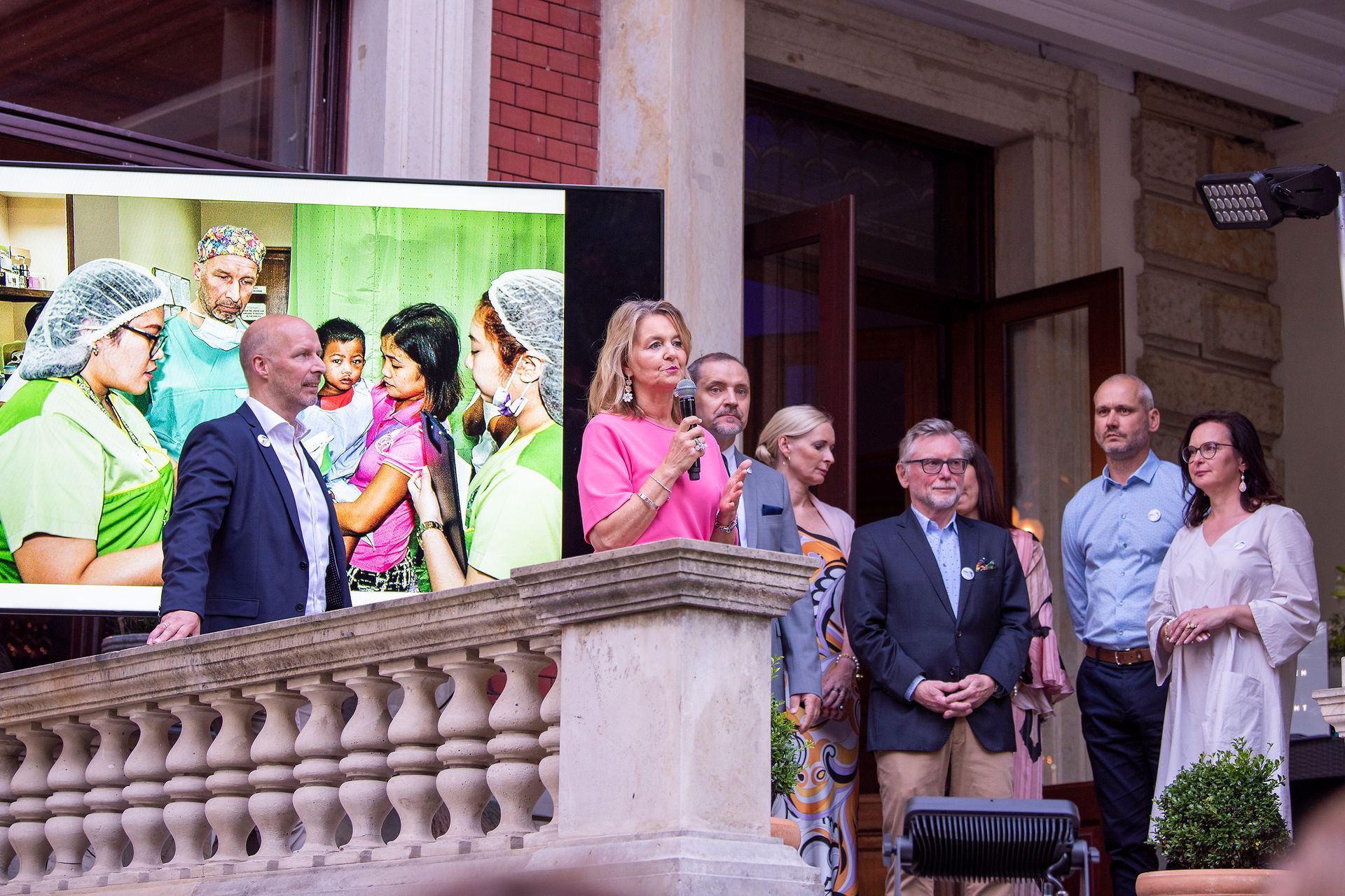 Eine Gruppe von Menschen steht auf einem Balkon vor einem großen Bildschirm mit einem Bild von Menschen darauf