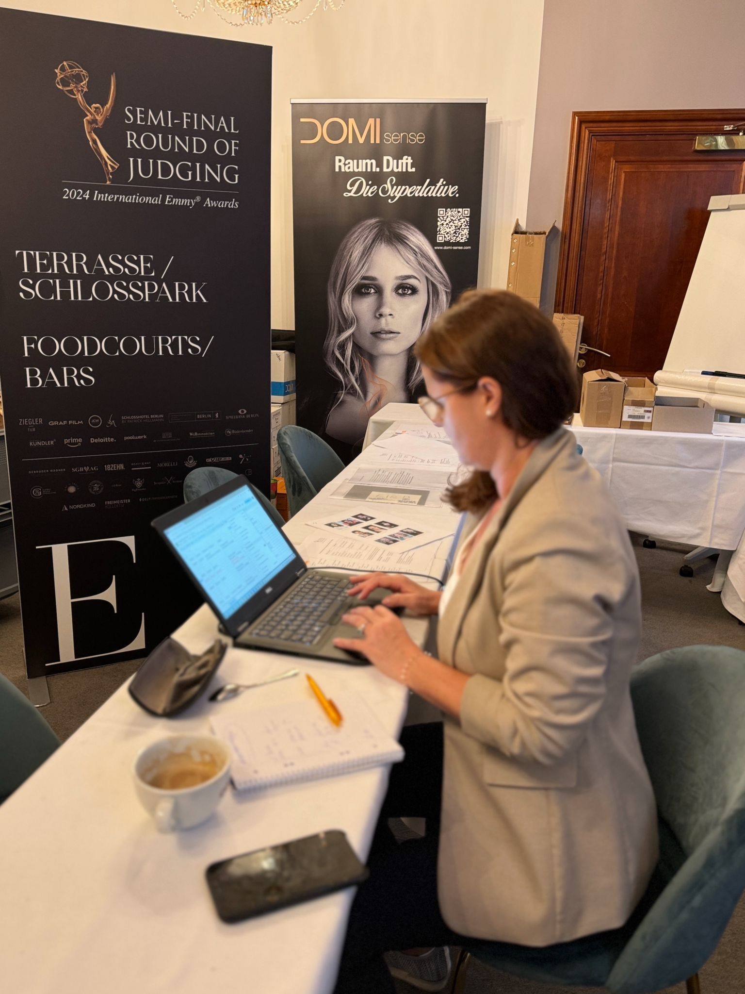 Eine Frau sitzt mit einem Laptop an einem Tisch vor einem Schild mit der Aufschrift „Domi“