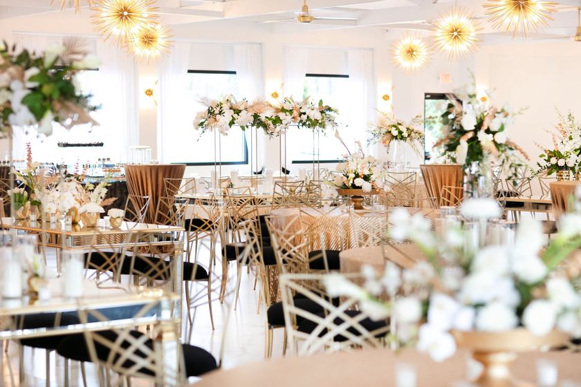 Revelry 675's reception room set up with 60-inch round tables featuring gold tablecloths, mirrored tables , gold chameleon chairs with black cushions, and white, green, and gold floral arrangements.
