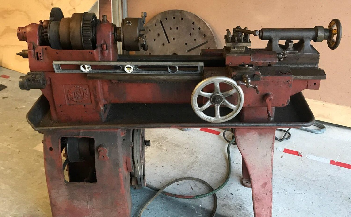 Red metal lathe on stand in a workshop, with attached chuck and handwheels.