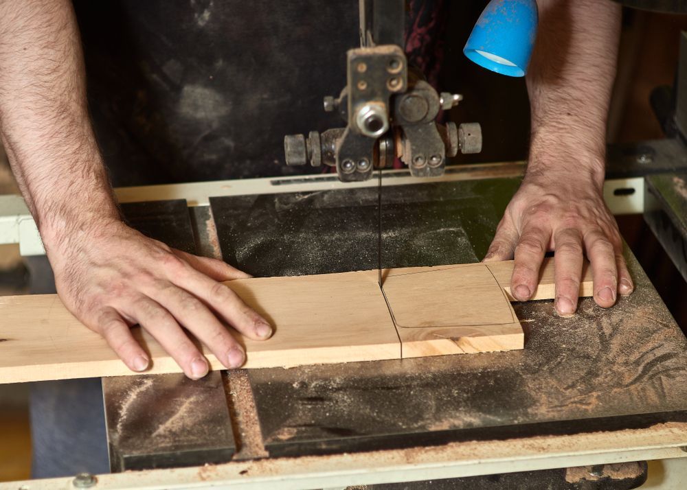 Hands guiding a wooden plank through a band saw to cut it. Saw is in a workshop setting.