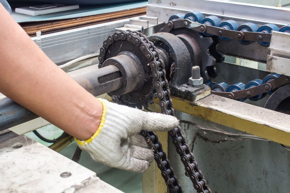 Gloved hand holding a chain, likely for machinery maintenance. Gears, rollers, and metal components are visible.