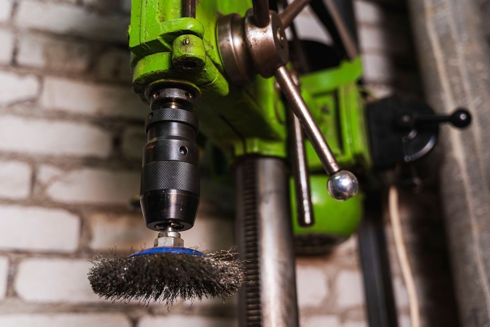 Green drill press with wire brush attachment, near a brick wall.