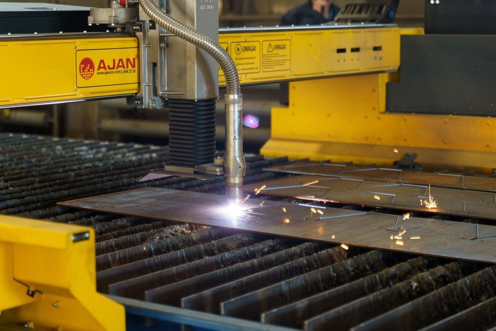 Plasma cutter cutting metal sheet in a factory setting. Sparks fly as the machine moves. Yellow machine arms.