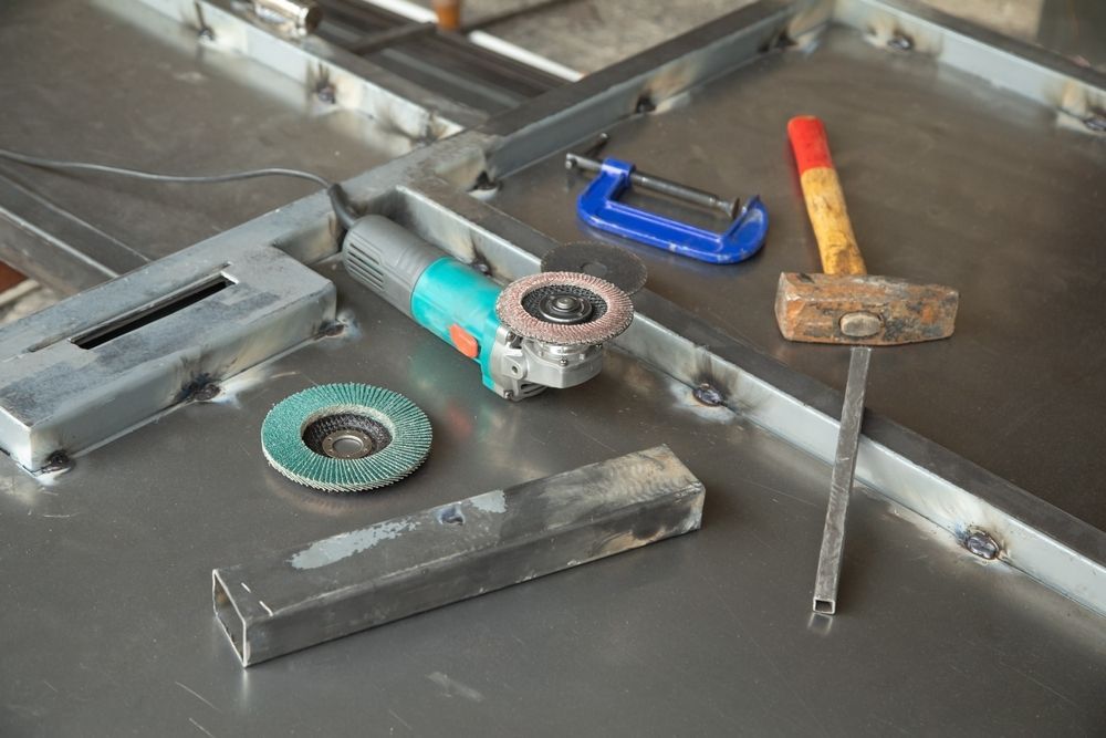 Metalworking tools: grinder, hammer, clamp, and metal pieces on a table.