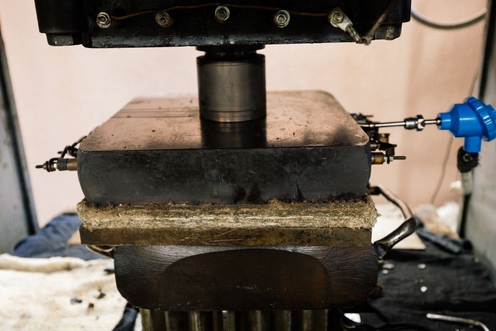 A metal press compressing a stack of materials, with a blue temperature sensor on the side.