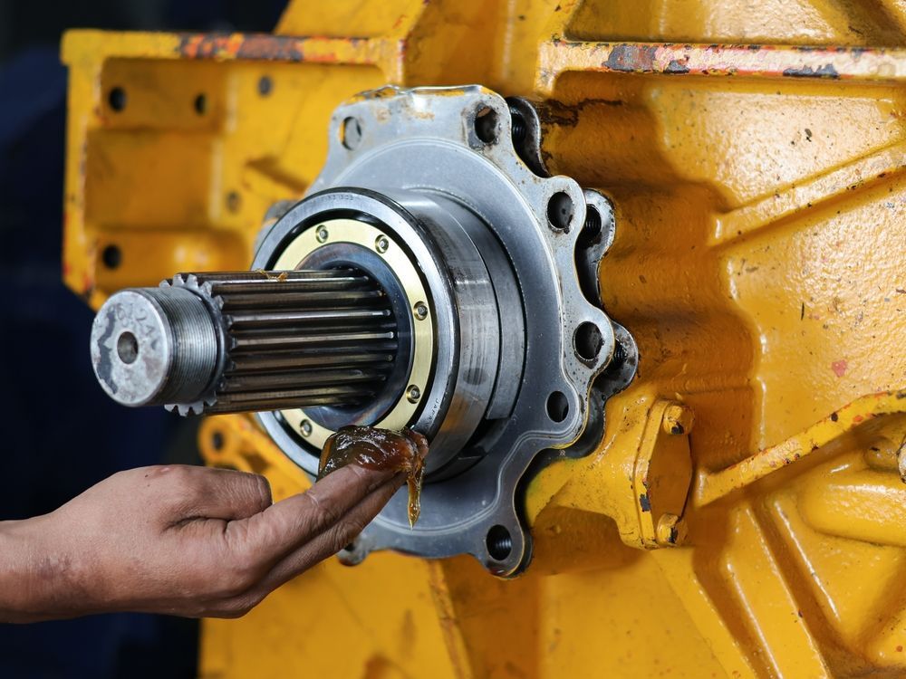 Hand applying grease to a metal shaft extending from yellow machinery, indoors.