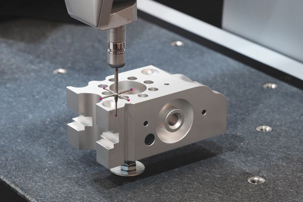 CMM machine inspecting a silver metal block with various holes and features on a granite table.