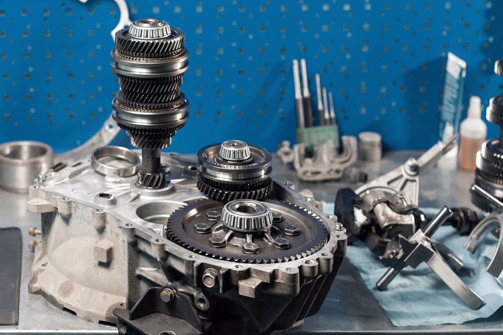 Disassembled car transmission parts on a workbench. Gears, bearings, and casing visible. Blue background.