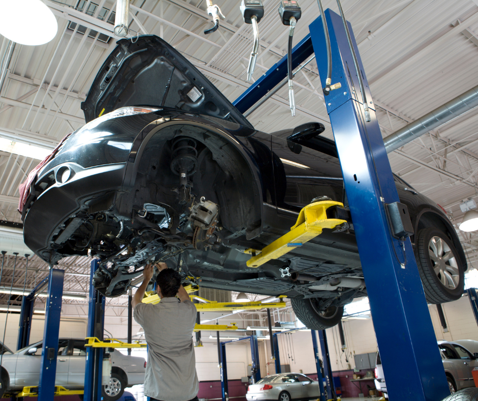 Car on a lift in a garage, mechanic working underneath. Black car, yellow lift arms, blue lift posts.