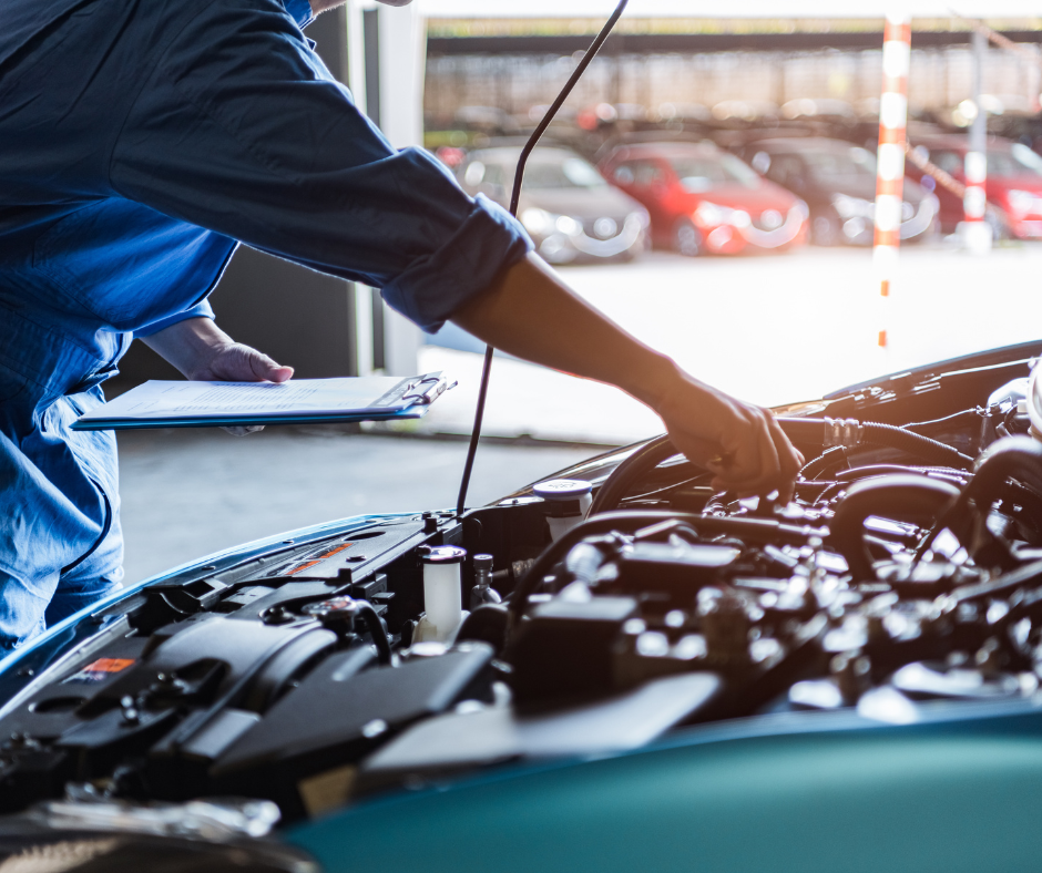 Mechanic in blue jumpsuit examining car engine with a clipboard in a garage.