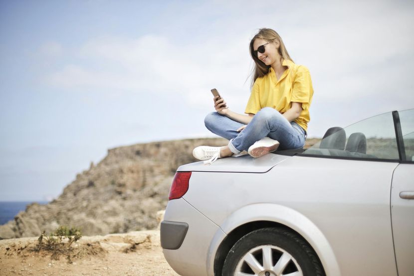 Woman in yellow shirt and jeans sits on a convertible car, using a phone, with a scenic view.