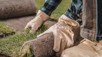 Gloved hands laying sod over soil in a grassy yard, with boots and jeans visible