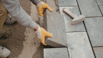 Worker laying gray paving stones beside a sandy base with a mallet nearby