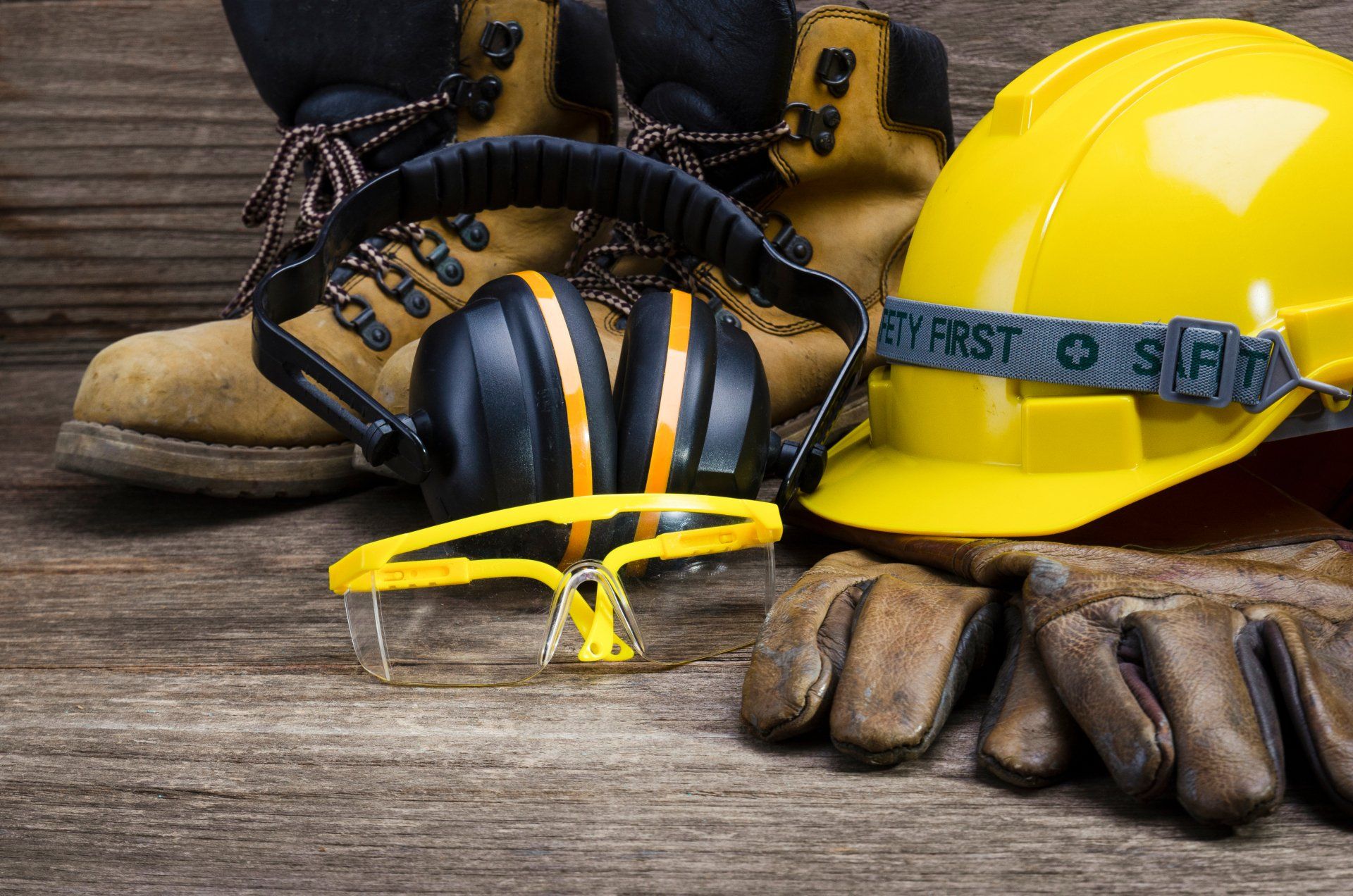 A hard hat , ear muffs , safety glasses , gloves and boots are on a wooden table.