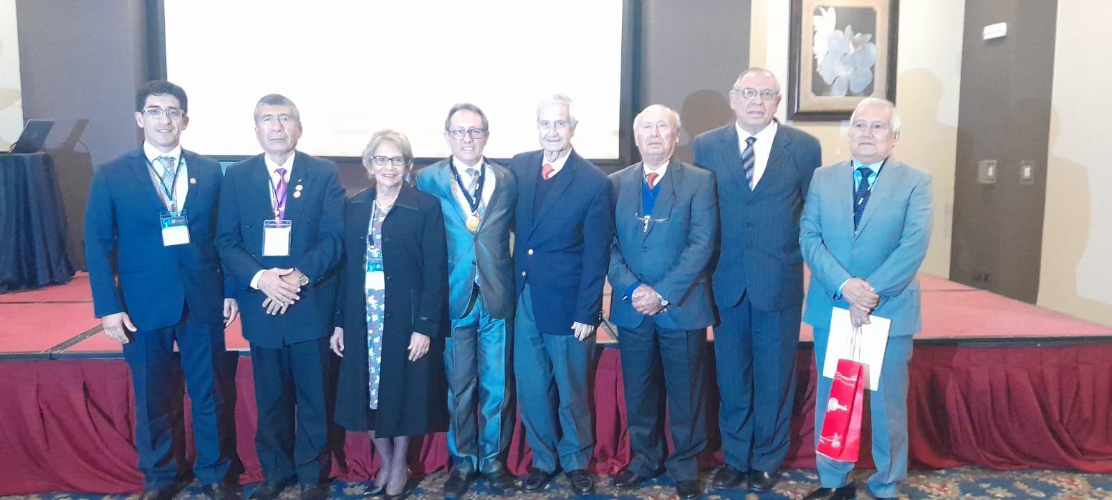 Grupo de personas de pie en un escenario durante un evento, algunas de ellas vestidas con trajes y cordones.