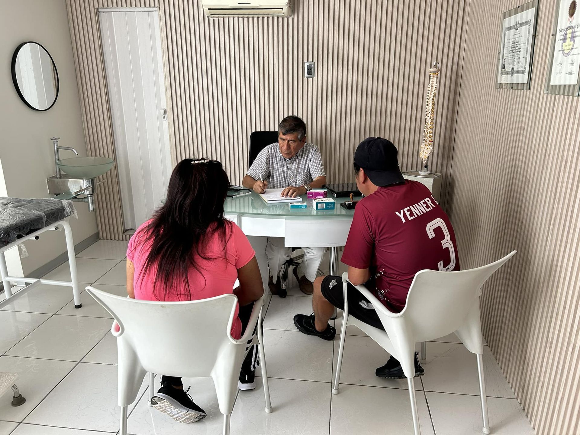 Médico consultando a dos pacientes en un consultorio. Mesa blanca, sillas y pared de rayas beige.