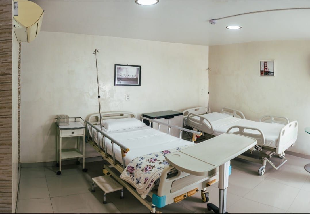 Habitación de hospital con dos camas, soporte para suero, mesita de noche pequeña e iluminación cenital.