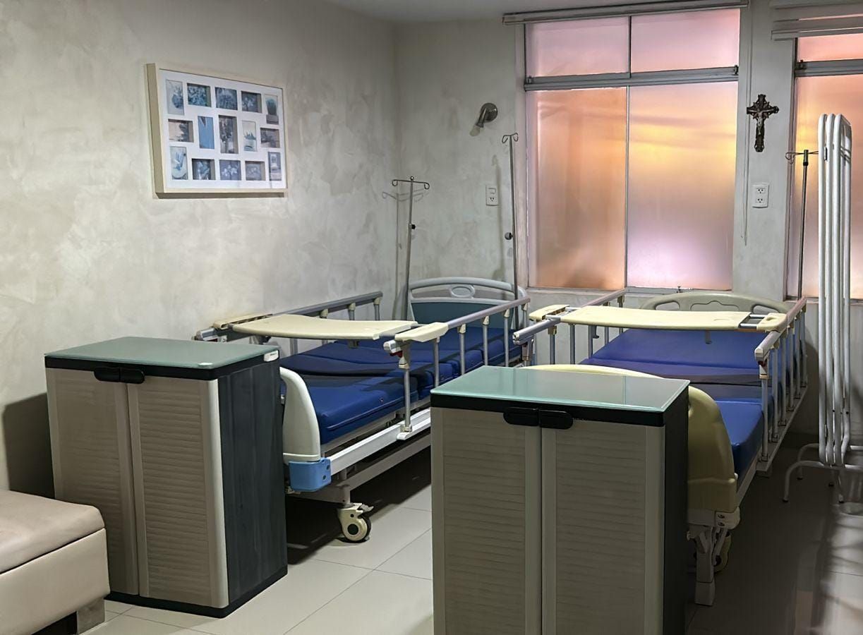 Dos camas de hospital en una habitación, con portasueros, armarios y una cruz en la pared. Luz tenue desde la ventana.