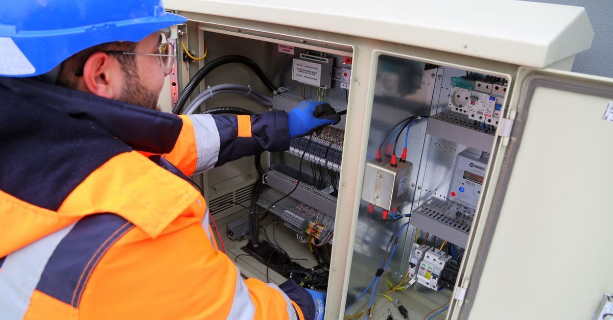 Técnico con chaleco de seguridad naranja y casco azul inspeccionando equipos eléctricos en un armario exterior abierto.