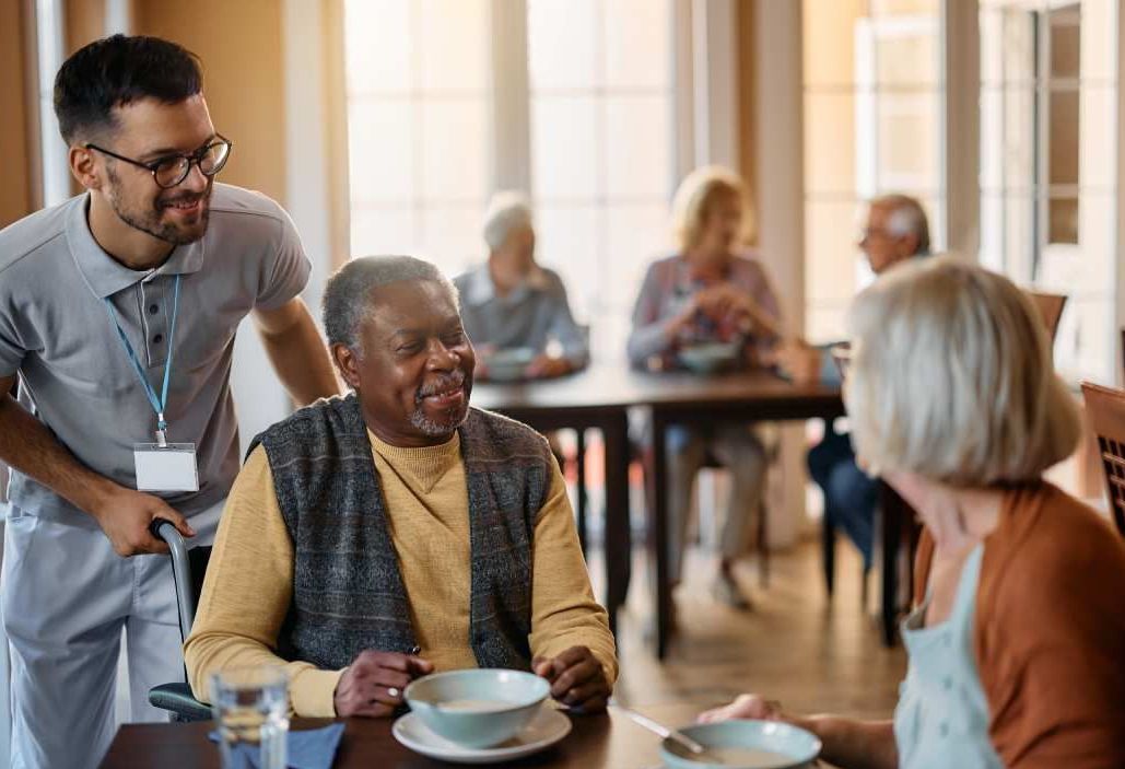 A man is serving food to an elderly man and woman in a nursing home.