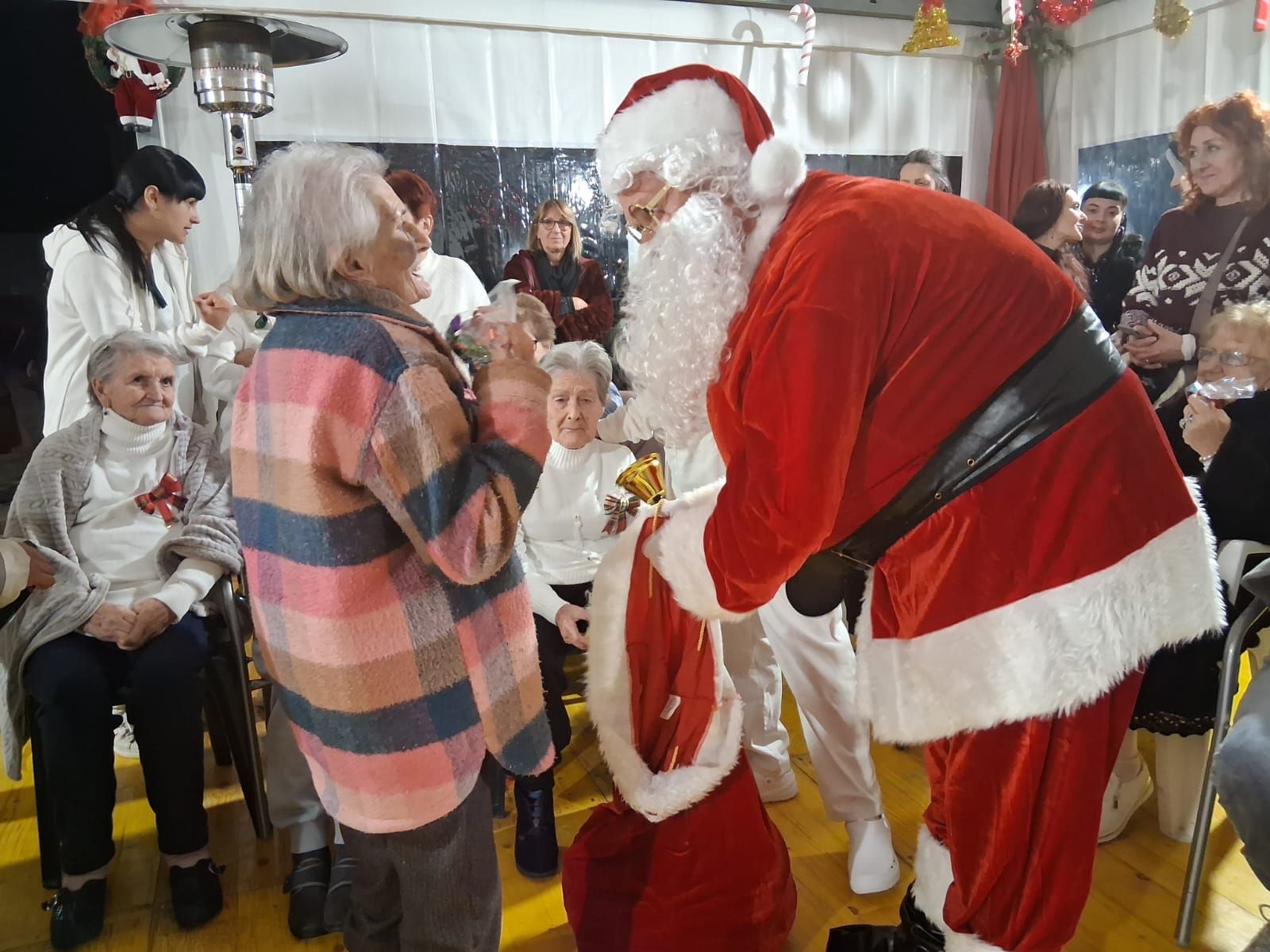 Babbo Natale in piedi in una stanza addobbata, porge un regalo dal suo sacco rosso a un anziano con una camicia a quadri.